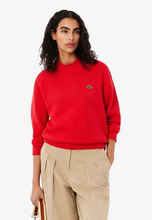Maglione rosso a maglia con scollatura rotonda a coste e maniche a sbuffo. Presenta un piccolo logo di coccodrillo verde sul petto. Abbinato a pantaloni beige.