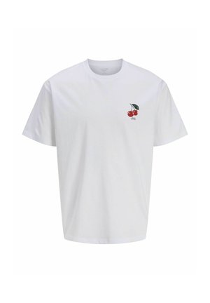 Garçon aux cheveux foncés portant un t-shirt blanc avec un motif cerise, un jean large bleu et des bottes couleur sable, debout devant un fond clair uni.