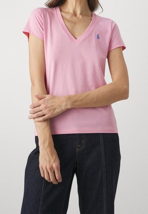 Kobieta ubrana w różową koszulkę z krótkim rękawem i dekoltem w kształcie litery V, z małym niebieskim logo na lewej piersi, w ciemnych dżinsach, ręka spoczywa na przeciwległym ramieniu.