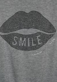 Grauer Stoff mit einem großen schwarzen Strassdesign in Lippenform, in dessen Mitte der Text "SMILE" steht, sowie einer handschriftlichen Akzentuierung darunter.