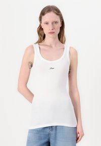 Camiseta sin mangas blanca de canalé con tirantes anchos, escote redondo y un pequeño logo negro en el centro. La tela parece suave y ajustada.