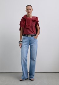 Chemise rouge à manches courtes avec un pull drapé sur les épaules, associée à un jean large bleu clair et une ceinture noire. Des sandales simples complètent le look.