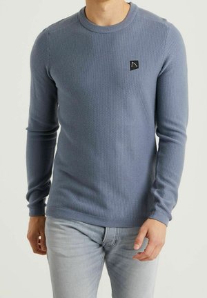 Hombre que lleva un suéter de cuello redondo de manga larga texturizado en azul polvo, con un pequeño logo negro en el lado izquierdo del pecho, combinado con jeans gris claro.