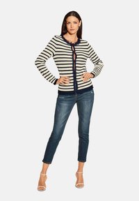 Gestreepte cardigan in marineblauw en crèmekleur, met een franjeszoom en knoopsluiting. Gecombineerd met donkere denimjeans en sandalen met hak.