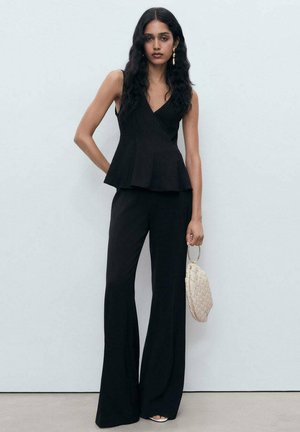 Crni peplum top s V-izrezom uparen s crnim širokim hlačama. Model drži tkanu torbicu svijetle boje. Glatke teksture i čiste linije.