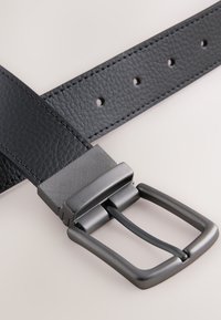 Zwarte leren riem met een gestructureerde donker metalen gesp; heeft meerdere gaatjes voor aanpassing en een gladde afwerking van de riem.