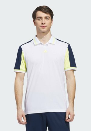 Junger Mann mit weißem Poloshirt mit marineblauen und gelben Ärmeln, steht vor einem schlichten hellen Hintergrund.