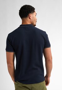 Námořnická polokošile s texturovanou tkaninou, krátkými rukávy a kontrastním límečkem. Nošena s olivově zelenými šortkami, pohled zezadu.