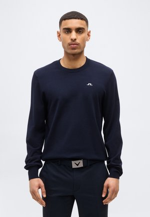 Hombre vistiendo un suéter azul marino con un pequeño logo blanco y pantalones oscuros con un cinturón que tiene una hebilla plateada, de pie contra un fondo liso.