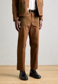 Brown cotton trousers with contrasting khaki patches, straight-leg design, and a textured finish. Paired with black lace-up shoes.