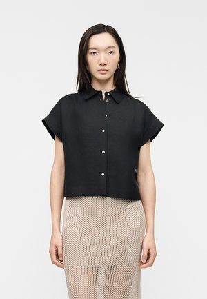 Kvinde iført en sort kortærmet skjorte med knapper og et beige net-nederdel, stående mod en ensfarvet lys baggrund.