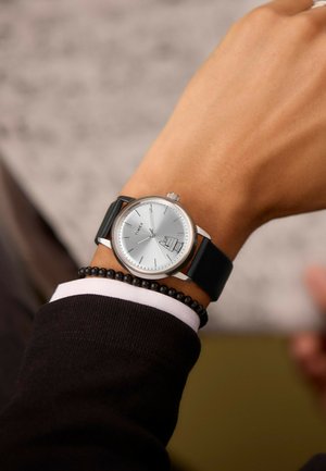Montre-bracelet Timex argentée avec un bracelet en cuir noir au poignet d'une personne, indiquant l'heure à 7h11, avec une petite icône de gâteau près du chiffre 4.