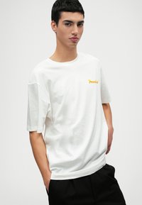 Camiseta de algodón blanca de corte holgado, con mangas cortas y escote redondo. Presenta un pequeño gráfico amarillo de "Paradise" en el pecho.