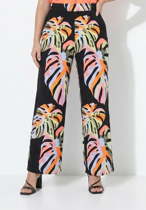 Vrouw die zwarte wijde broek draagt met een meerkleurige grote tropische bladprint en zwarte sandalen met hakken.