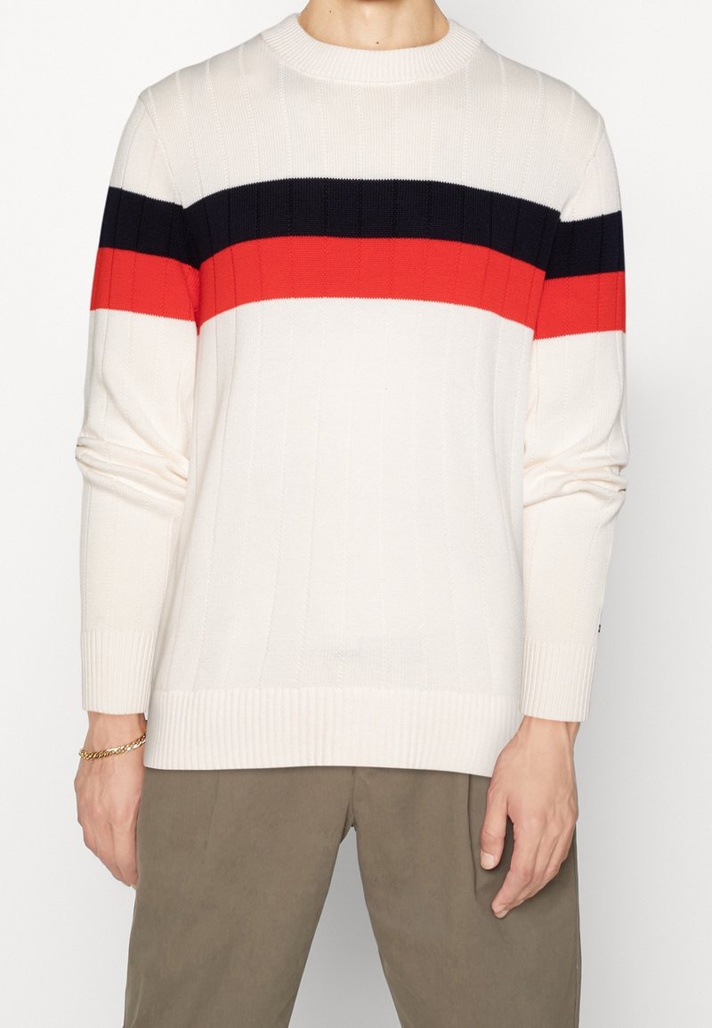 Personne portant un pull en tricot blanc avec des rayures horizontales noires et rouges sur la poitrine et un pantalon kaki, debout les bras détendus.