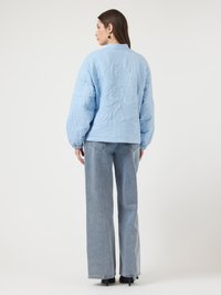 Γυναίκα με πλάτη φοράει ανοιχτό μπλε μπουφάν με καπιτονέ ύφανση, ανοιχτόχρωμο τζιν παντελόνι με φαρδιά γραμμή, μαύρα παπούτσια με τακούνι και μαύρα σκουλαρίκια, πάνω σε λευκό φόντο.