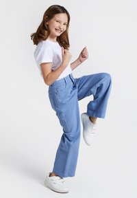 Jasnoniebieskie spodnie o szerokich nogawkach z tylnymi kieszeniami, w połączeniu z białą koszulką i białymi trampkami, w pozycji stojącej z jedną nogą uniesioną.