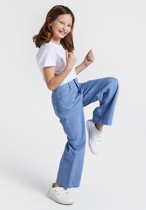 Jasnoniebieskie spodnie o szerokich nogawkach z tylnymi kieszeniami, w połączeniu z białą koszulką i białymi trampkami, w pozycji stojącej z jedną nogą uniesioną.