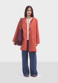 Giacca leggera arancione con cappuccio e bottoni, abbinata a una maglietta bianca, pantaloni larghi a righe blu e una borsa bordeaux.