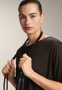 Czarny krótki top z gładką teksturą. Modelka trzyma cienki, czarny sznur przymocowany do ciemnego obiektu blisko swojej klatki piersiowej na neutralnym tle.