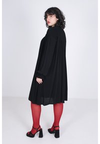 Zwarte lange jurk met smokdetail bij de halslijn, vloeiende design, gecombineerd met rode panty’s en zwarte hoge hakken.