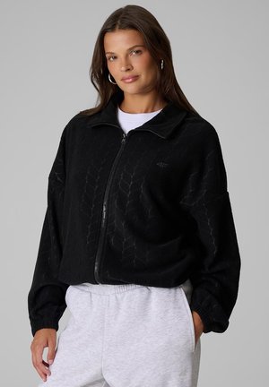 Femme aux longs cheveux bruns portant une veste zippée noire à motifs et un pantalon de jogging gris clair, la main dans une poche.