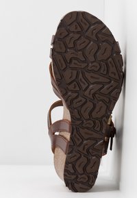 Sandale en cuir marron avec semelle extérieure en caoutchouc texturé, semelle intermédiaire en liège et bride à boucle, positionnée debout sur un fond blanc.