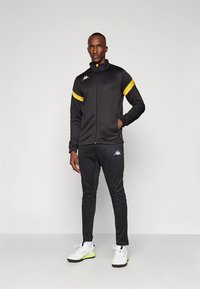 Črna atletska trenirka z rumenimi poudarki na ramenih. Jakna ima zadrgo; hlače so zožene. Čevlji so beli z zelenimi detajli.