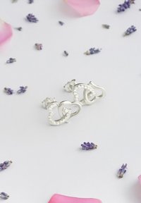 Boucles d'oreilles en argent en forme de boucles entrelacées, ornées de strass transparents, sur un fond clair avec des pétales de lavande et de rose.