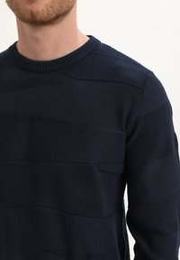 Merinopusero, jossa on vaakasuoria tekstuuriraitoja, ribbireunus kauluksessa ja tiukka istuvuus. Pehmeä, keskikokoinen kangas.