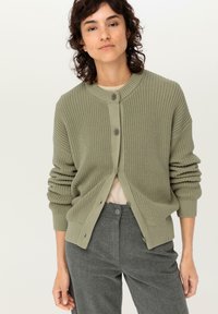 Grön stickad cardigan med rund halsringning, knappstängningar och uppvikta ärmslut. Kombineras med grå byxor och en beige topp under.