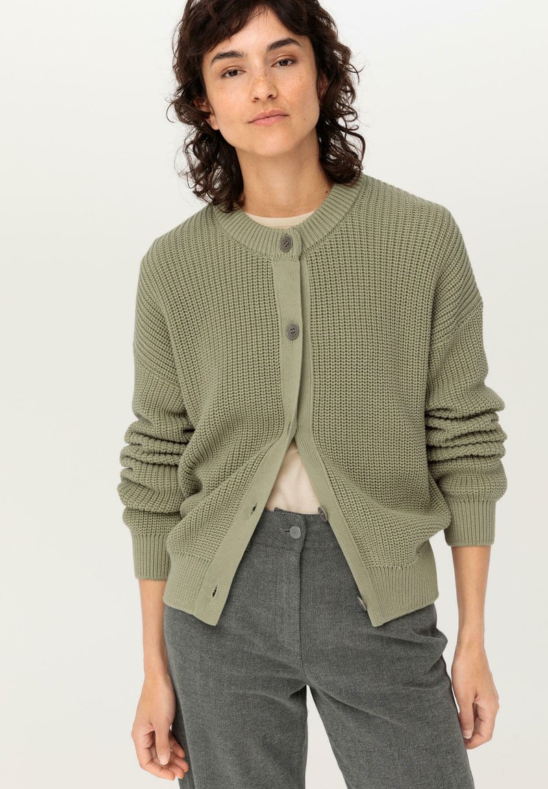 Grön stickad cardigan med rund halsringning, knappstängningar och uppvikta ärmslut. Kombineras med grå byxor och en beige topp under.