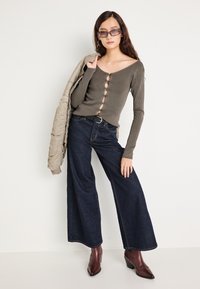 Γυναίκα που φοράει γκρι ριμπ πουλόβερ με κουμπιά, φαρδιές σκούρες μπλε τζιν παντελόνες, μπορντό μπότες, γυαλιά ηλίου και κρατάει μια μπεζ ζακέτα στον ώμο.