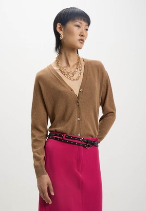 Cardigan marrone a maglia con scollo a V e bottoni, abbinato a una cintura nera con borchie su una gonna rosa brillante. Il tessuto testurizzato contrasta i colori.