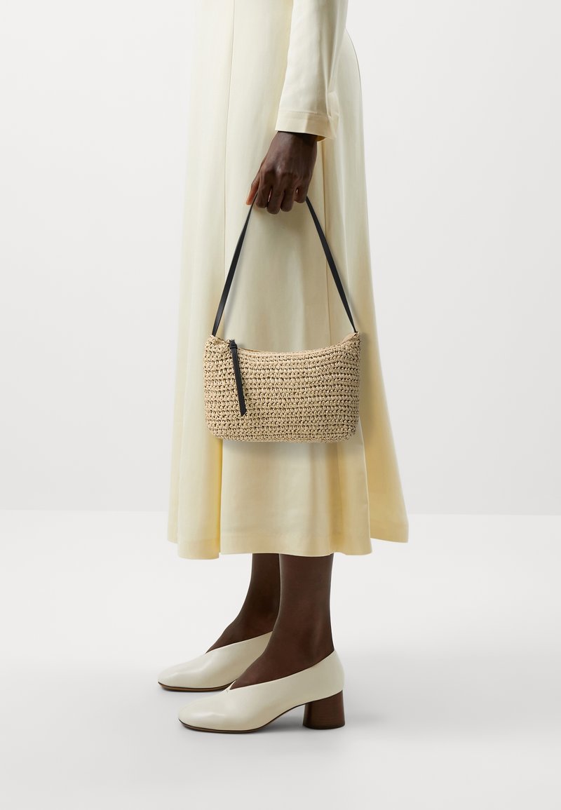 Tkaná béžová kabelka s černou rukojetí, zipovým uzavřením a strukturovaným povrchem. V kombinaci s dlouhými, světlými žlutými šaty a krémově bílými botami na blokovém podpatku.