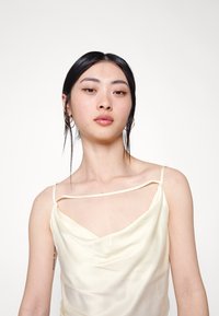 Camisola de seda em amarelo pálido com decote drapeado, alças finas e um brilho subtil. Apresenta um corte solto e fluido.