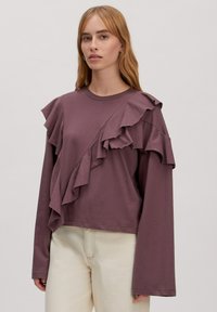 Lila Langarmoberteil mit geschichteten Rüschen auf einer Seite, weichem Baumwollmaterial und Rundhalsausschnitt. Getragen mit hellen Hosen.