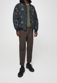 Bomberka w wzór kamuflażu w odcieniach zieleni i czerni, zestawiona z oliwkowym swetrem z okrągłym dekoltem i brązowymi spodniami, dopełniona czarnymi sneakersami.