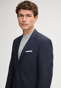 Marynarski blazer w wzór w paski z klapami notch i zapięciem na jeden guzik, w połączeniu z jasnoszarym golfem i białą chusteczką w kieszeni.