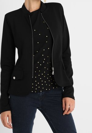 Femme portant un blazer noir ajusté zippé sur une chemise noire à petits pois dorés et un jean bleu foncé, mains détendues le long du corps.