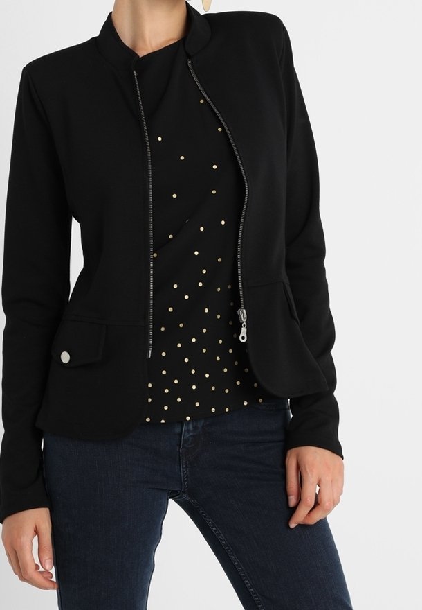 Femme portant un blazer noir ajusté zippé sur une chemise noire à petits pois dorés et un jean bleu foncé, mains détendues le long du corps.