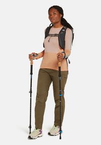 Donna che indossa una camicia a maniche lunghe pesca, pantaloni oliva, scarpe da trekking e uno zaino nero, mentre tiene in mano due bastoni da trekking e guarda dritto davanti a sé.