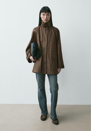 Žena v hnědém kabátu s vysokým límcem, modrých džínách a hnědých botách, držící černou tašku na jednobarevném světlém pozadí.