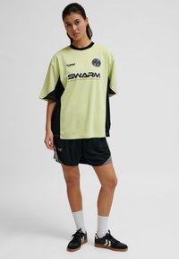 Lys grøn og sort sports trøje med korte ærmer, der har et logo og tekst på forsiden. Sammen med sorte shorts og sorte sko.