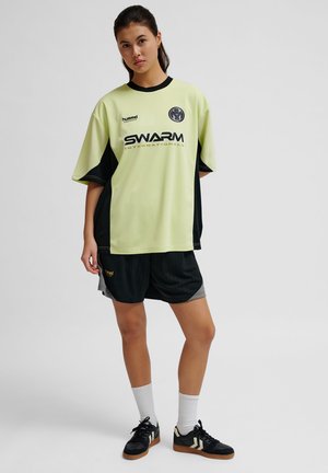 Lichtgroen en zwart sportshirt met korte mouwen, voorzien van een logo en tekst aan de voorkant. Gecombineerd met zwarte shorts en zwarte schoenen.