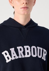 Moleton azul-marinho com cordão, apresentando o texto bordado em branco "BARBOUR" no peito. Tecido suave e corte descontraído.