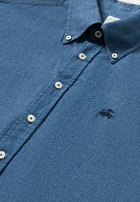 Overhemd met knopen in vervaagd blauw, gemaakt van zachte stof. Heeft een korte kraag, witte knopen en een klein marineblauw geborduurd logo.