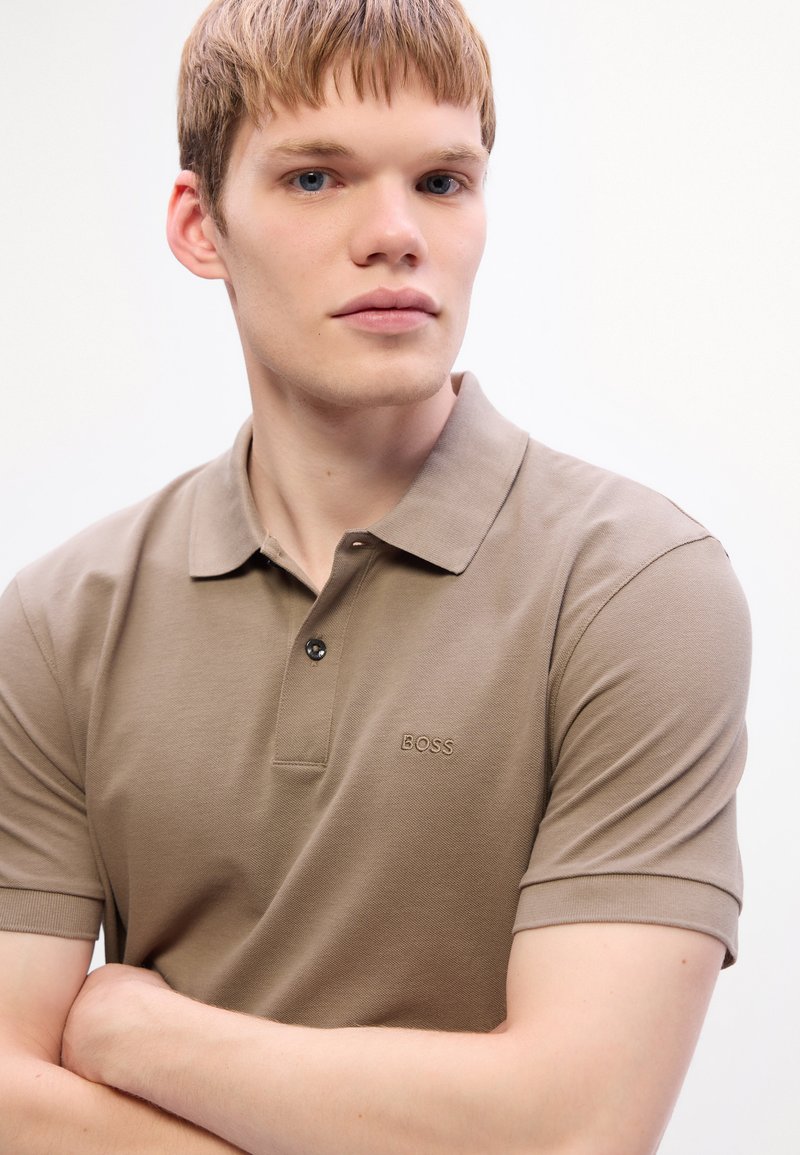 Jeune homme avec des cheveux courts châtain clair portant un polo beige BOSS, les bras croisés, regardant légèrement sur le côté devant un fond blanc.