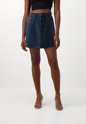 Woman wearing a dark blue denim mini skirt with small white studs, a black cropped top, and clear strap high-heeled sandals.