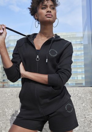 Felpa con zip nera realizzata in tessuto testurizzato, con logo sul petto, abbinata a pantaloni corti neri coordinati e con tirazip in metallo circolare.